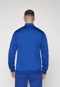 Blaues Sportjacke mit hochgeschlossenem Kragen, langen Ärmeln und gerippten Bündchen. Mit roten Akzenten an den Seiten. Glatte Textur, figurnahes Design.