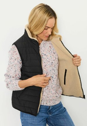Frau zeigt das weiche beige Fleecefutter ihrer schwarzen gesteppte Weste über einem gepunkteten Pullover und blauen Jeans.