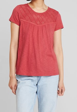 Vrouw draagt een rood topje met korte mouwen en een gebreid kapmouwdetail en lichtblauwe spijkerbroek tegen een effen achtergrond.