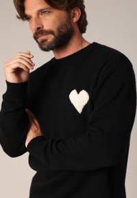 Zwarte trui van zachte stof met een wit hartontwerp op de linkerborst. Geribde manchetten en halslijn. Relaxte pasvorm.