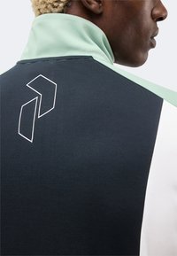 Άτομο που φοράει μαύρο και ανοιχτοπράσινο μπουφάν με ψηλό γιακά και λευκό γεωμετρικό λογότυπο στο πίσω μέρος, κοντά στον ώμο.