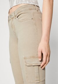 Beiga cargobyxor med ficka på locket, bärs av en person med synlig mage i en ljus mönstrad topp, med höger hand vilande nära låret.