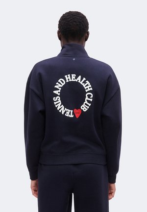 Person iført en mørkeblå jakke med "Tennis and Health Club" i et cirkulært design og et rødt hjerte med "Borg" på ryggen.