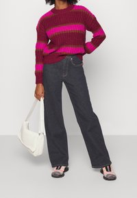 Stickad tröja i bordeaux med rosa och olivfärgade ränder, i kombination med mörk denimjeans. Tillbehören inkluderar en vit vävd väska och flerfärgade sneakers.