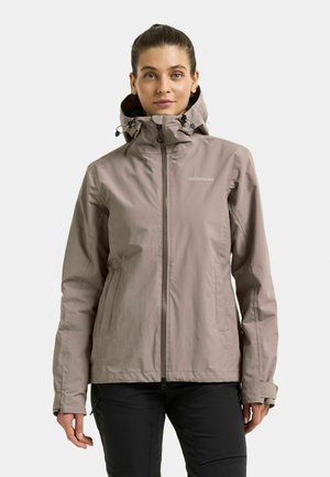 Kvinde vendt fremad, iført en taupe hættejakke med lynlås og "Didriksons"-logo på brystet samt sorte bukser, mod en ensfarvet baggrund.