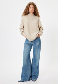Pull en maille beige avec un col haut, associé à un jean à jambes larges bleu clair. Le tissu semble doux avec une finition texturée.