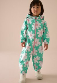 Tag-glo-grüner Blumenregenanzug mit großen weißen Gänseblümchen und pinken Strahlen, Reißverschluss, elastischen Bündchen und Kapuze. Weiße Stiefel runden den Look ab.