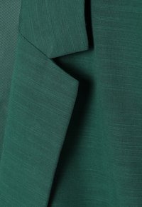 Gros plan d’un tissu vert avec texture de tissage visible, montrant un matériau plié et superposé avec des lignes de couture nettes.