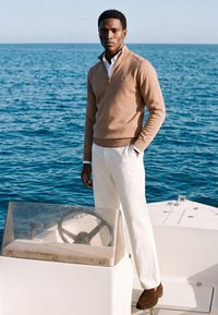 Homme portant un pull beige et un pantalon blanc, debout sur un bateau, une main dans la poche, avec la mer en arrière-plan.