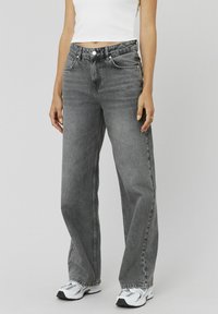Jeans gris à jambes larges avec une teinte délavée, design à cinq poches, fermeture par bouton et coutures visibles. Portés avec un top blanc ajusté et des baskets.