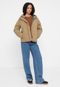 Giubotto puffer marrone con cappuccio, caratterizzato da un design trapuntato, chiusure a scatto e tasche laterali. Abbinato a jeans blu a gamba larga e scarpe nere.