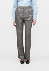 Pantalon gris plissé avec une coupe ajustée, présentant une texture de tissu lisse et une taille mi-haute, associé à des chaussures noires élégantes.