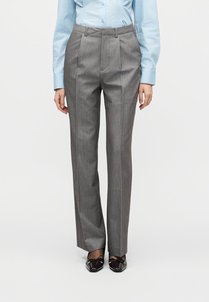 Pantalon gris plissé avec une coupe ajustée, présentant une texture de tissu lisse et une taille mi-haute, associé à des chaussures noires élégantes.