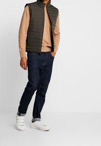 Gilet trapuntato verde scuro sopra un maglione beige chiaro, abbinato a jeans blu scuro e sneakers bianche con cinturini in Velcro. Design semplice e funzionale.
