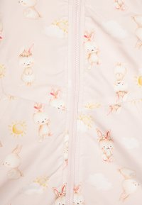 Tissu rose clair avec un imprimé fantaisiste de lapins, de soleils et de nuages. Détail comprenant une fermeture éclair centrale et une surface douce et texturée.