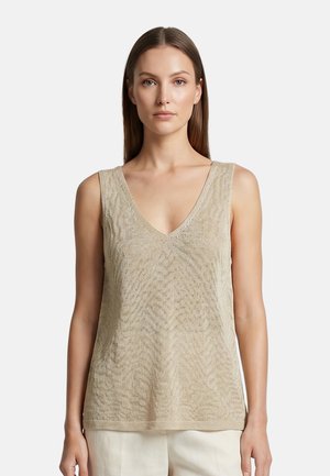Donna con capelli castani lisci indossa un top beige senza maniche in maglia con scollo a V e pantaloni chiari su uno sfondo semplice.