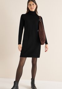 Schwarzes gestricktes Rollkragenkleid mit langen Ärmeln, gerader Schnitt, kombiniert mit einer braunen Wildleder-Schultertasche und schwarzen Schnürstiefeln.