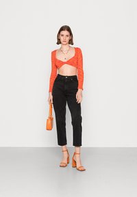 Suéter encurtado vermelho-laranja com mangas longas, combinado com calças pretas de cintura alta, sandálias de salto laranja e uma mala laranja.