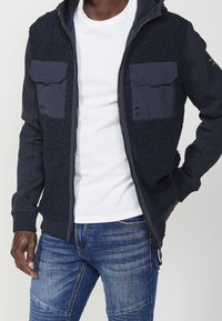 Marinblå zip-up jacka med fluffig textur på framsidan, två bröstfickor och ribbade mudd. Bärs över en vit t-shirt och blå jeans.