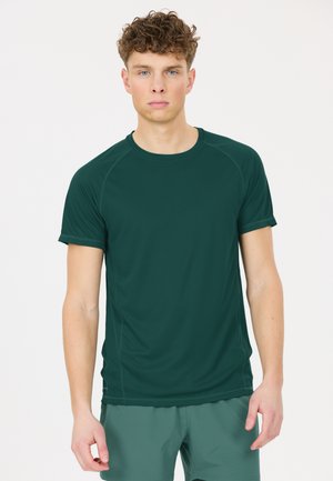 Jonge man met kort krullend haar draagt een donkergroen sportshirt met korte mouwen en een groene korte broek en staat tegen een effen lichte achtergrond.