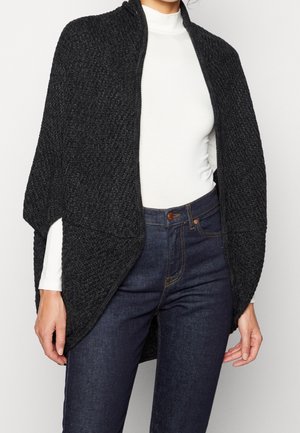 Femme portant un châle tricoté noir texturé ouvert sur le devant, par-dessus un haut blanc à manches longues et un jean bleu foncé, debout devant un fond clair.