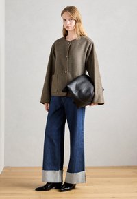Kvinde iført en oversized brun jakke, brede bukser med manchetter i blå denim, sorte hælsko, holder en stor foldet sort lædertaske, står på trægulv.