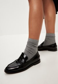 Svarta lackskinnsloafers med två dekorativa remmar, i kombination med grå ankelsockor; slät textur med en elegant och klassisk design.