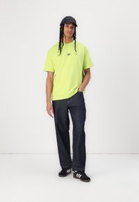 Neon green short-sleeve t-shirt with small logo, dark blue straight-leg jeans, and black cap with braided hair, standing in a neutral background.