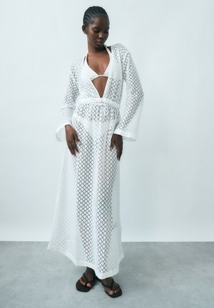 Kvinde iført hvid bikini og lang, gennemsigtig hvid overdel med geometriske mønstre, stående på gråt gulv mod en ensfarvet hvid væg.