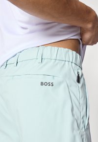 Svetlomodré športové šortky s elastickým pásom, bočným vreckom na zips a sivým logom "BOSS" na pravej stehne. Hladká textúra látky.