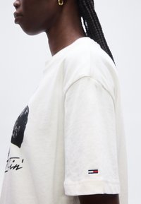 Μαύρη γυναίκα με πλεξούδες που φοράει μια λευκή φαρδιά μπλούζα με μικρό λογότυπο σημαίας της Tommy Hilfiger στο μανίκι.