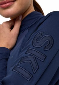 Navyblaue Langarmoberteil mit strukturiertem Stoff und einem gestickten "SKI"-Logo in dunklerem Blau auf dem linken Ärmel. Nahaufnahme.