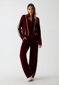 Bordeaux velours trainingspak met een ritsjas met witte strepen en bijpassende broek met een elastische tailleband en zijzakken.