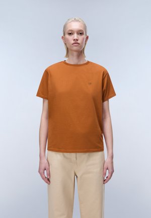 Kobieta z krótkimi blond włosami ubrana w gładką, spalonych pomarańczowy t-shirt i beżowe spodnie, stoi na jasnoszarym tle.