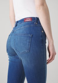 Mujer vistiendo jeans azules de MAX&Co. y una camiseta sin mangas blanca, mostrada desde la cintura hacia abajo en un ligero ángulo lateral.