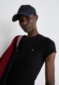 Námornícka čiapka, čierne priliehavé tričko s malým logom a červená/námornícka varsity bunda so pruhovanými akcentmi, vystavené na svetlom pozadí.