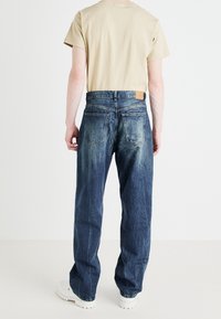 Vaqueros azules de mezclilla con acabado desgastado, pierna recta y bolsillos traseros, combinados con una camiseta beige y zapatillas blancas gruesas.