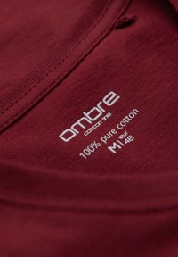 Etiqueta del cuello de una camiseta de algodón burdeos, con texto blanco que dice "línea de algodón ombre 100% algodón puro M EUR 48" sobre una superficie de tela suave.