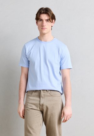 Jeune homme aux cheveux bruns portant une chemise à manches courtes bleu clair et un pantalon beige sur un fond uni gris clair.