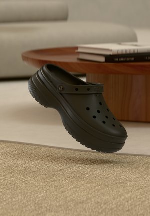 Sort gummiklogg med perforert design, forhøyede kanter og bakre stropp. Teksturert såle for grep. Flytende over en vevd overflate.