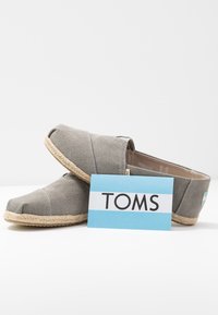 Espadrilles en toile grise avec un embout arrondi, une semelle en corde de jute et une étiquette en tissu bleu et blanc avec le logo. Modèle à dos ouvert.