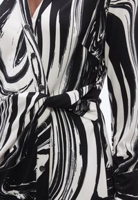 Svart-hvitt kjole med et virvlende, abstrakt mønster, knapper foran og en knytebelte i midjen. Glatt stofftekstur.