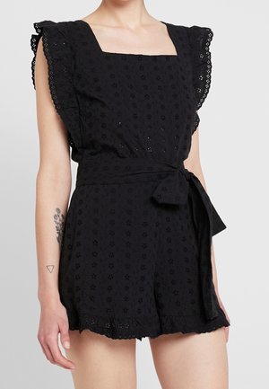 Schwarzer Eyelet-Overall mit quadratischem Ausschnitt, gerafften Kappenärmeln, gebundenem Taillengürtel und gewelltem Saum, getragen von einer stehenden Person mit Armtattoos.