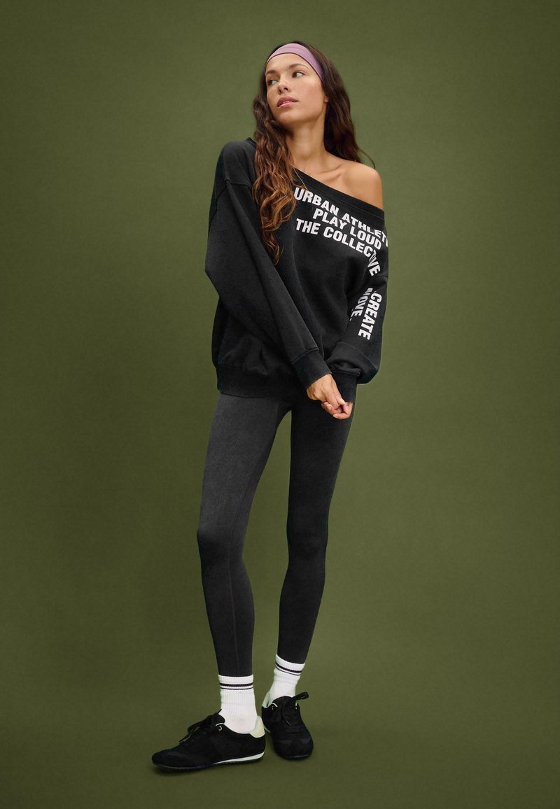 Felpa nera oversize con testo bianco, abbinata a leggings scuri e sneakers nere. L'outfit presenta accenti sportivi e un cerchietto.