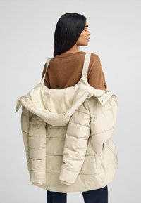 Chaqueta acolchada color crema con diseño de rombos, gran capucha y correas ajustables. Textura suave, corte oversized, detalle de marca minimalista.