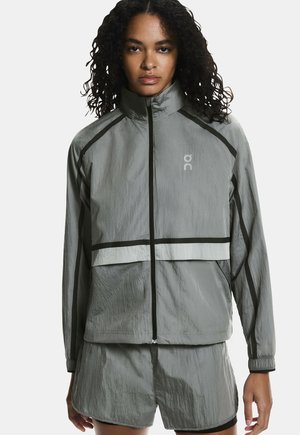 Mlada ženska, oblečena v sivo lahko vetrovko z zadrgo in ujemajoče kratke hlače, stoji ob enobarvni beli podlagi.