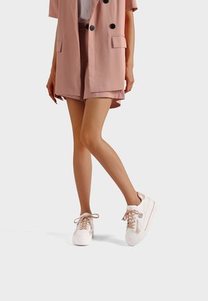 Donna con pantaloncini rosa chiaro e blazer, indossando scarpe da ginnastica bianche, in piedi con le gambe incrociate contro uno sfondo semplice.