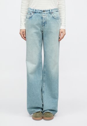 Jean large bleu clair avec finition délavée, cinq poches et détails de couture subtils. Porté avec des chaussures vertes et un haut à rayures.