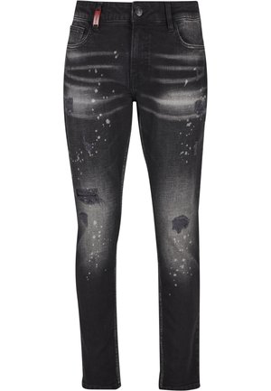 Homme debout de profil, portant un jean noir délavé avec des éclaboussures de peinture et des baskets blanches, beiges et noires.