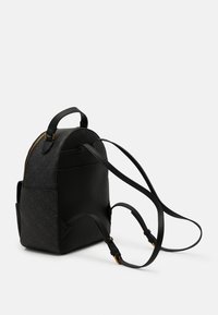 Sac à dos compact noir avec deux bretelles réglables, une poignée supérieure, des poches latérales et une fermeture éclair dorée sur un fond clair uni.